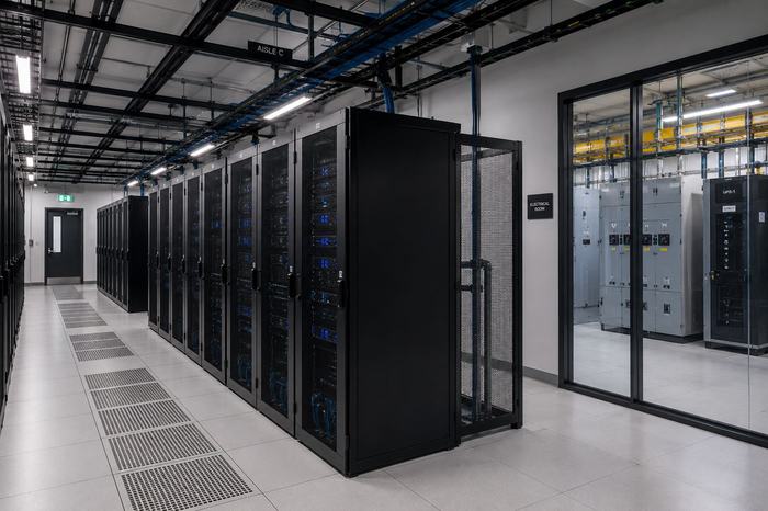 Pasillo de centro de datos con piso técnico perforado, bandejas portacables y sala eléctrica.